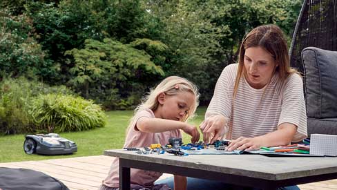 Frau und Kind entspannen im Garten, während Rasenroboter arbeitet