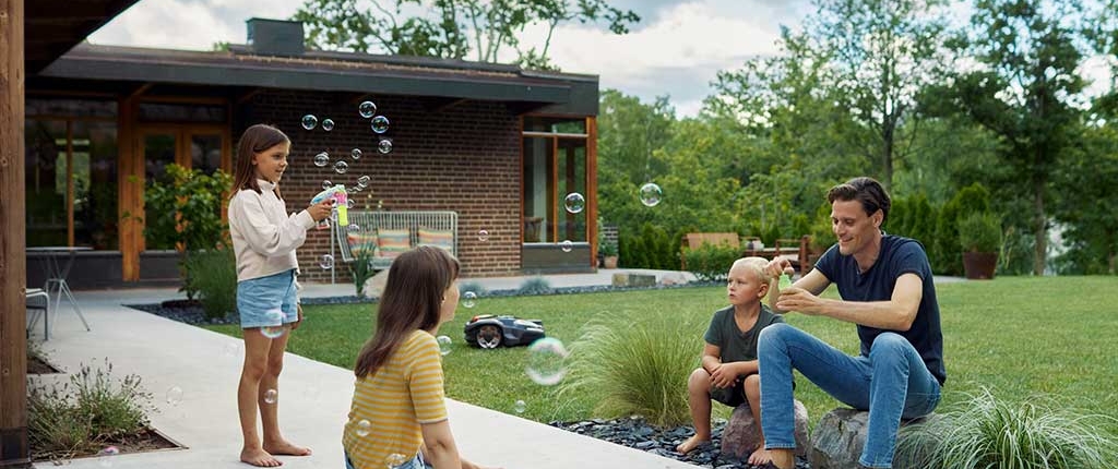 Vater spielt mit Kindern auf der Terrasse während der Husqvarna-Rasenrobotor die Wiese mäht.