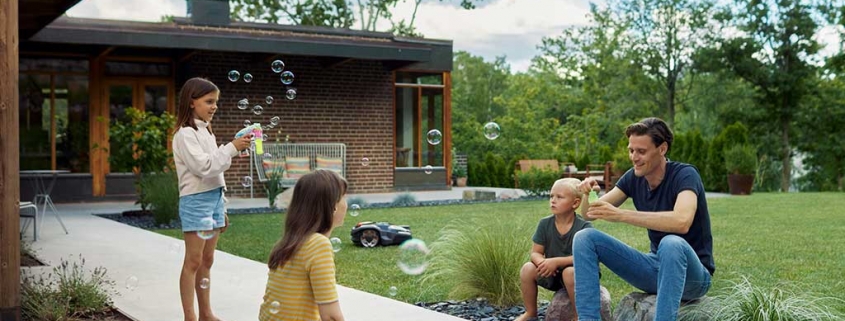 Vater spielt mit Kindern auf der Terrasse während der Husqvarna-Rasenrobotor die Wiese mäht.