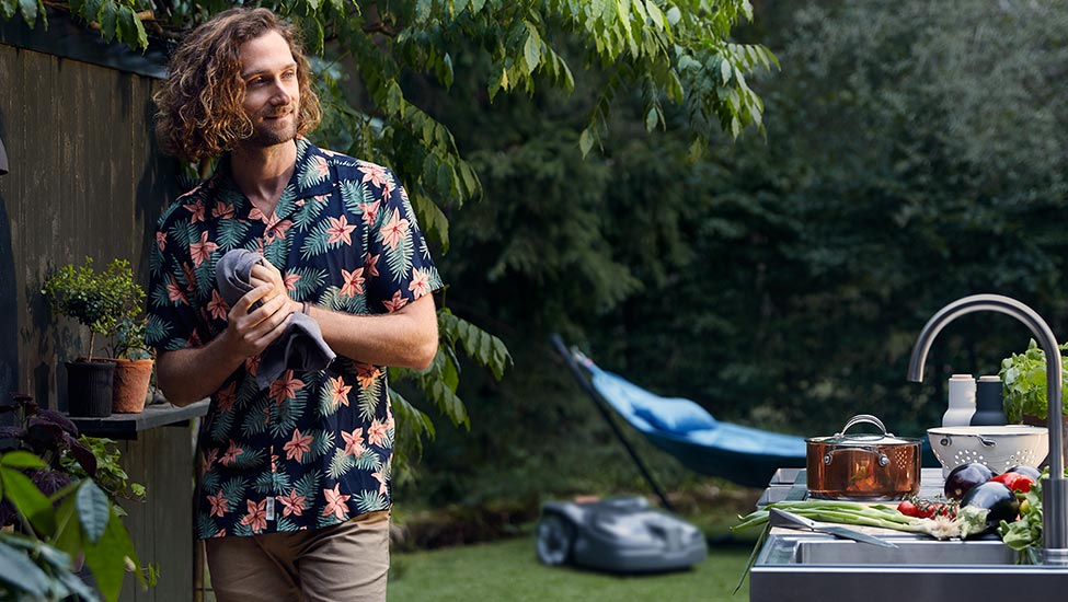 Mann grillt im Garten, Husqvarna Rasenroboter im Hintergrund