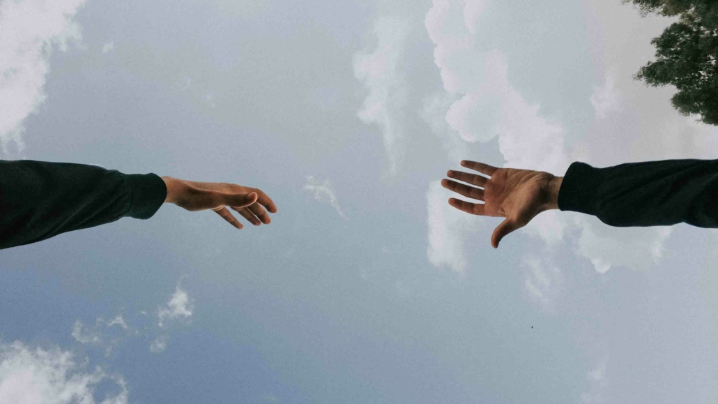 Zwei Hände, die versuchen, sich zu berühren vor bewölktem Himmel - Wir helfen Dir und nehmen Dich bei der Hand.