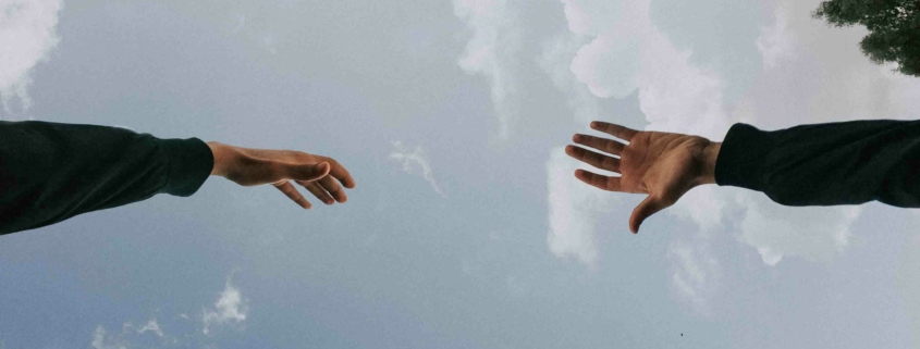 Zwei Hände, die versuchen, sich zu berühren vor bewölktem Himmel - Wir helfen Dir und nehmen Dich bei der Hand.